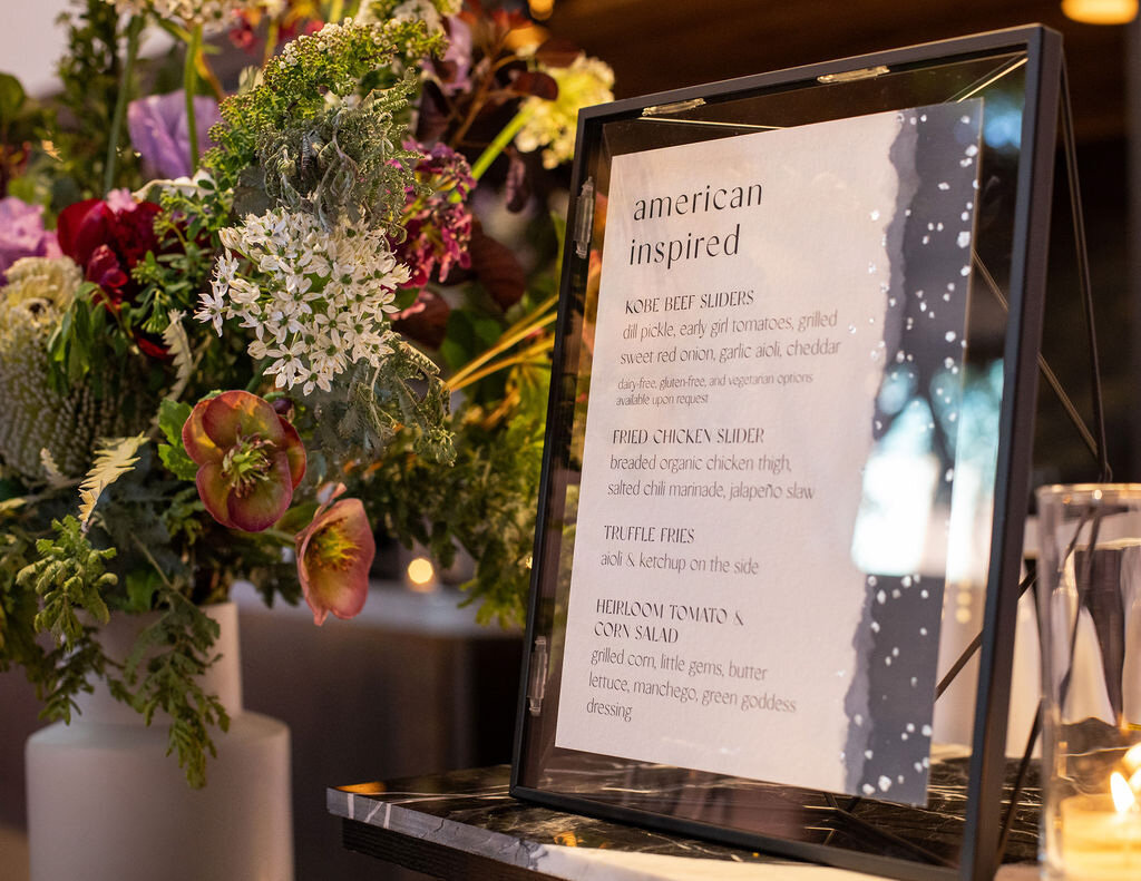 Menu of wedding reception food on a table next to a dark bouquet of flowers.